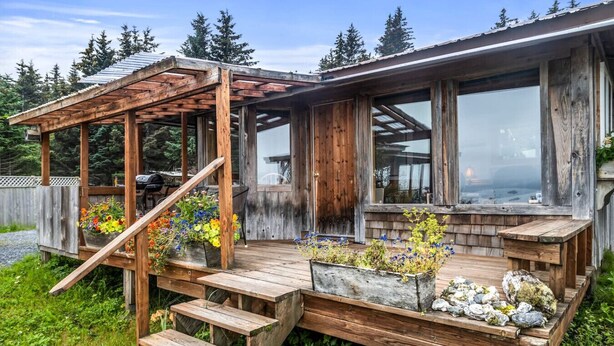 A wooden deck with steps leading to a covered area, wooden planters with flowers, and a view of the surrounding forest. - Rustic Roots Seaside Cardinal Cabin (Seward)