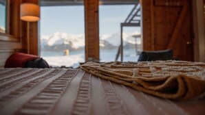 A wooden cabin room with a bed, a window, and a view of snow-capped mountains. - Rustic Roots Seaside Cardinal Cabin (Seward)
