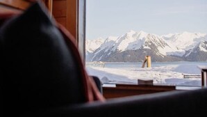 A cabin interior with a large window offering a view of snow-covered mountains and a lake. - Rustic Roots Seaside Cardinal Cabin (Seward)