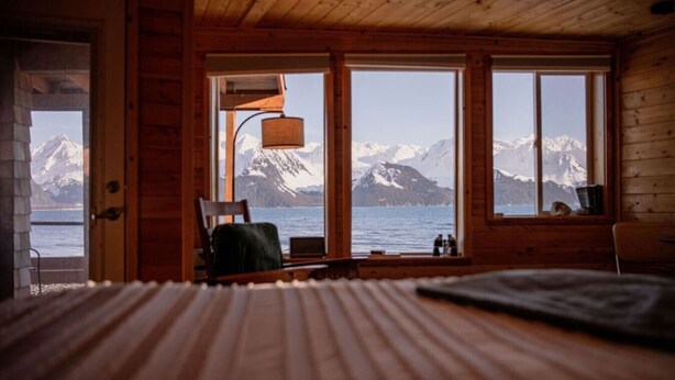 A wooden cabin room with a bed, a chair, and a view of mountains and water. - Rustic Roots Seaside Indigo Cabin (Seward)