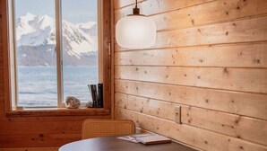 A wooden cabin interior with a round table, two chairs, a hanging light fixture, and a window with a view of snow-capped mountains. - Rustic Roots Seaside Indigo Cabin (Seward)