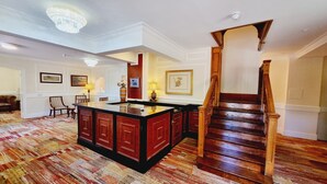 Een ruime woonkamer met een grote houten trap, een centraal eiland met kastjes, en een kamerbreed tapijt met patroon.