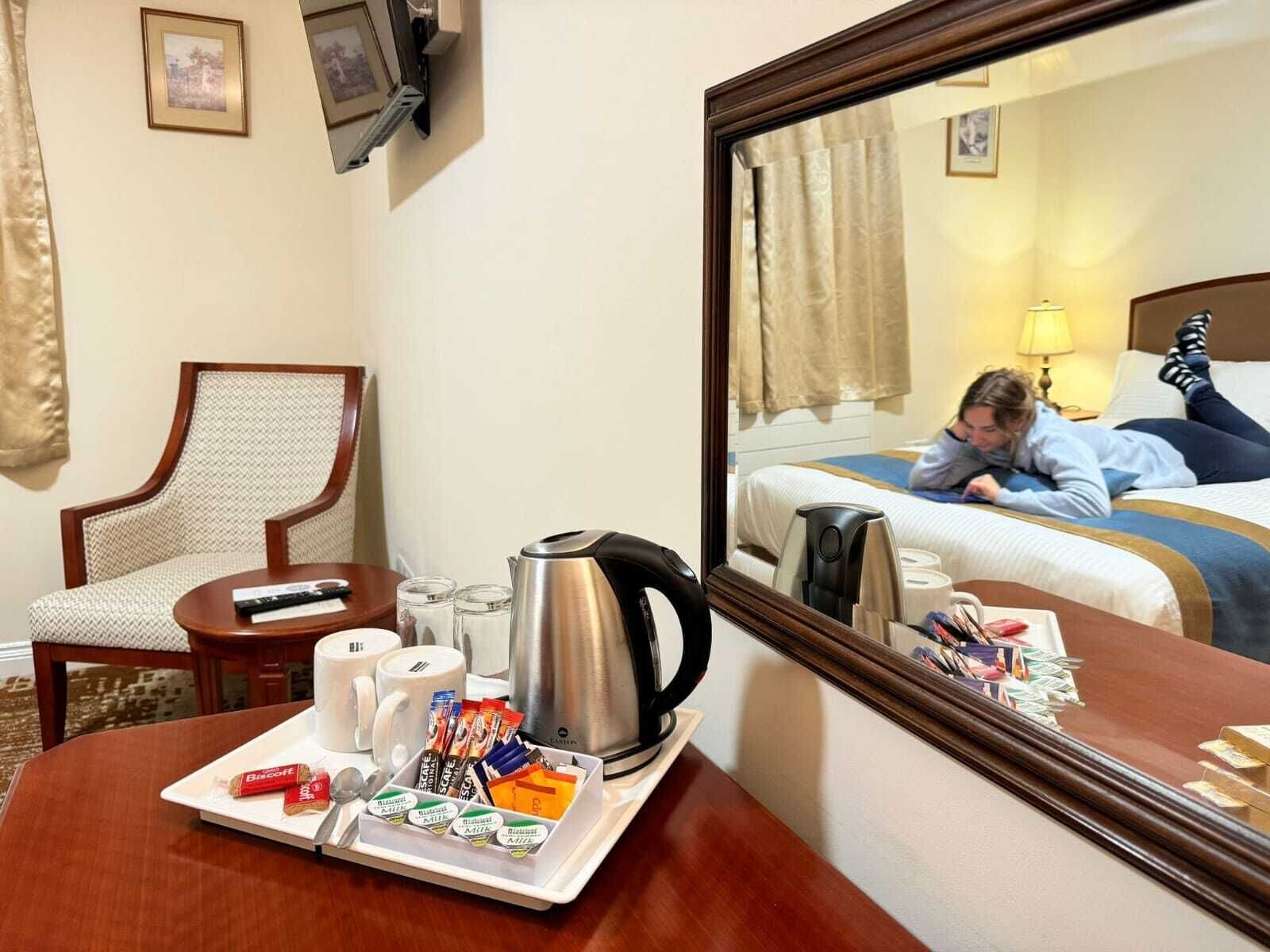 A hotel room with a bed, a bedside table with a kettle, cups, and a tray with snacks.