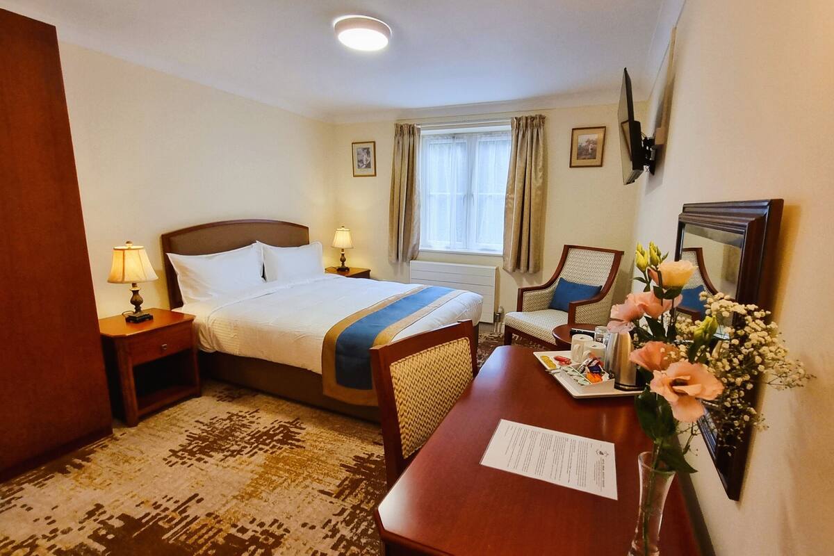 Hotellihuone, jossa on sänky, yöpöydät, työpöytä, tuoli, ikkuna verhoilla, peili ja maljakko, jossa on kukkia.