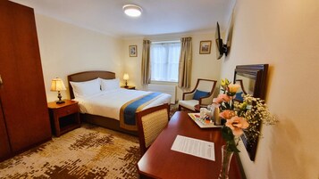 Habitación de hotel con cama, mesitas de noche, escritorio, silla, ventana con cortinas, espejo y un jarrón con flores.