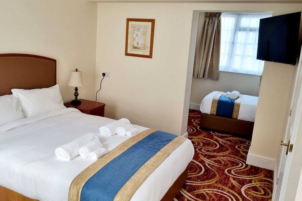 Hotellihuoneessa on suuri sänky, yöpöytä lampun kanssa, pieni pöytä pyyhkeille, taulu seinällä, seinälle asennettu televisio sekä toinen sänky viereisessä huoneessa.