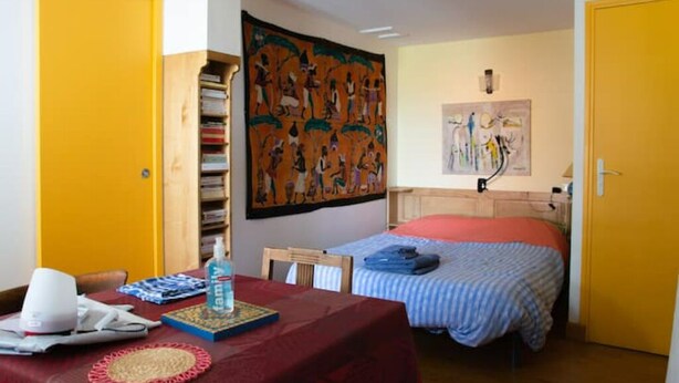 A room with a bed, a table with a red tablecloth, a bookshelf, and a large tapestry on the wall. - Studio Calme Chaleureux (Brive-la-Gaillarde)