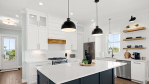 A modern kitchen with white cabinets, a large island, stainless steel appliances, pendant lighting, and wooden shelves.