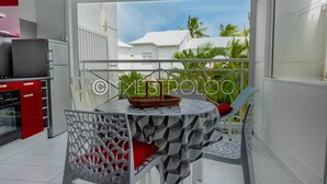 A modern kitchen with white cabinets and a red appliance, a dining area with a round table and chairs, and a balcony with a view of palm trees and a white building. - L'oasis du quai (Saint-François)
