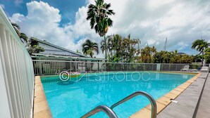 A swimming pool with clear blue water, surrounded by a white fence and palm trees. - L'oasis du quai (Saint-François)