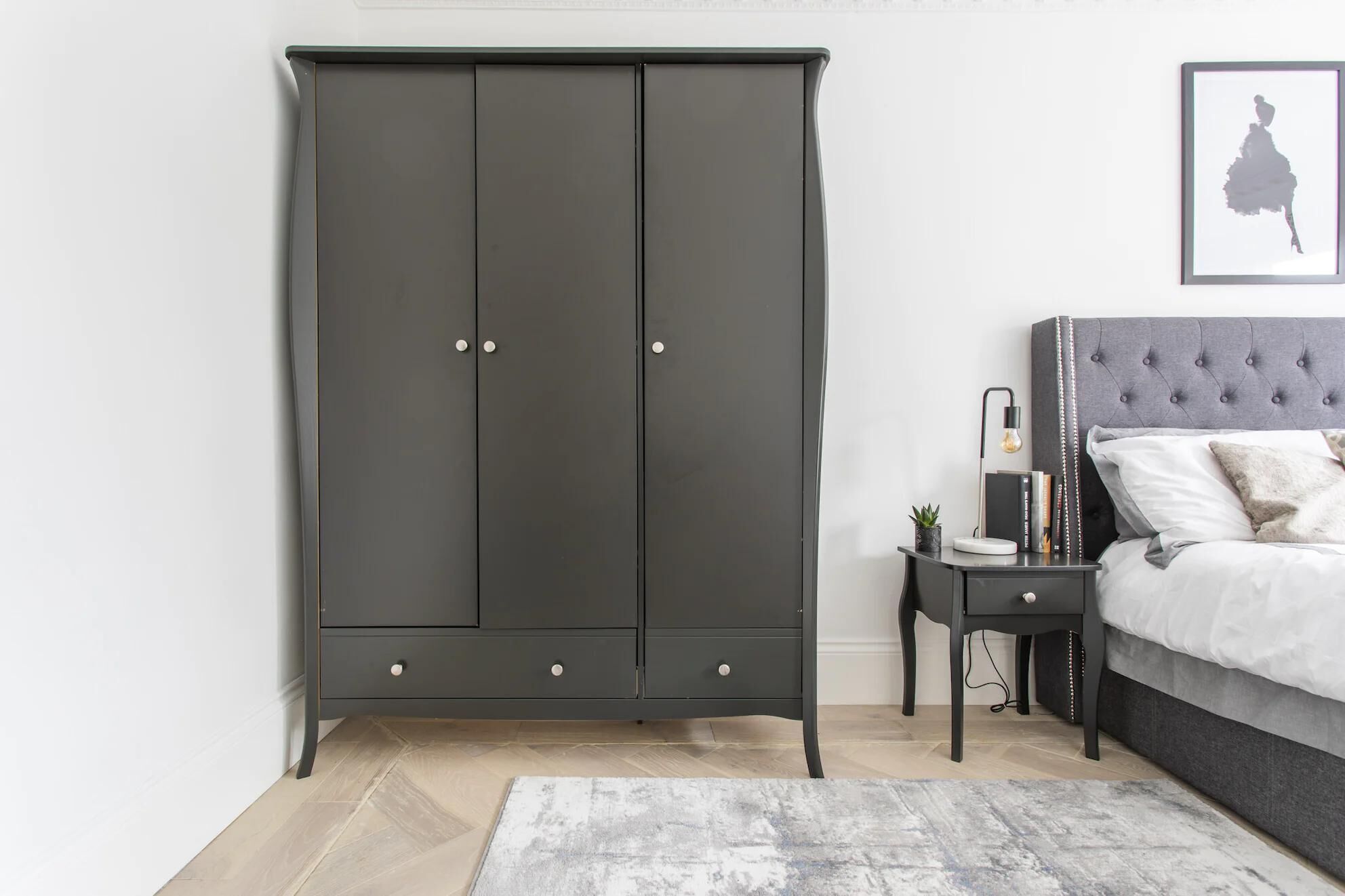 A bedroom with a large wardrobe, a bedside table with a lamp, and a bed with a grey headboard.