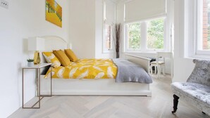 Une chambre lumineuse avec un lit, une table de chevet, une lampe, une chaise et de grandes fenêtres.