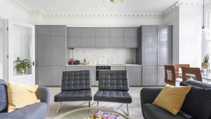 Un salon moderne avec deux canapés en cuir noir, une table basse en verre et un coin cuisine doté de meubles gris et d’un dosseret en marbre.
