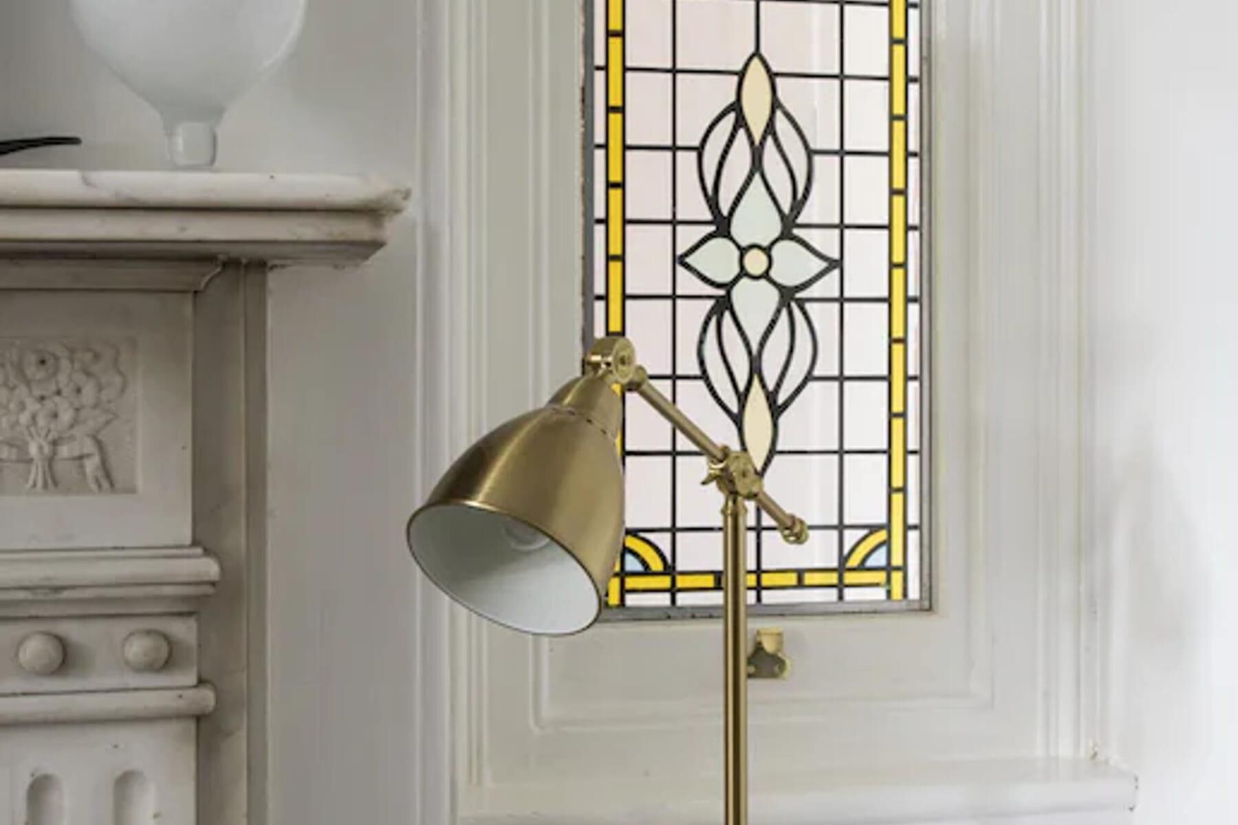 A brass floor lamp with a white shade positioned in front of a white wall with a decorative window featuring a geometric pattern.