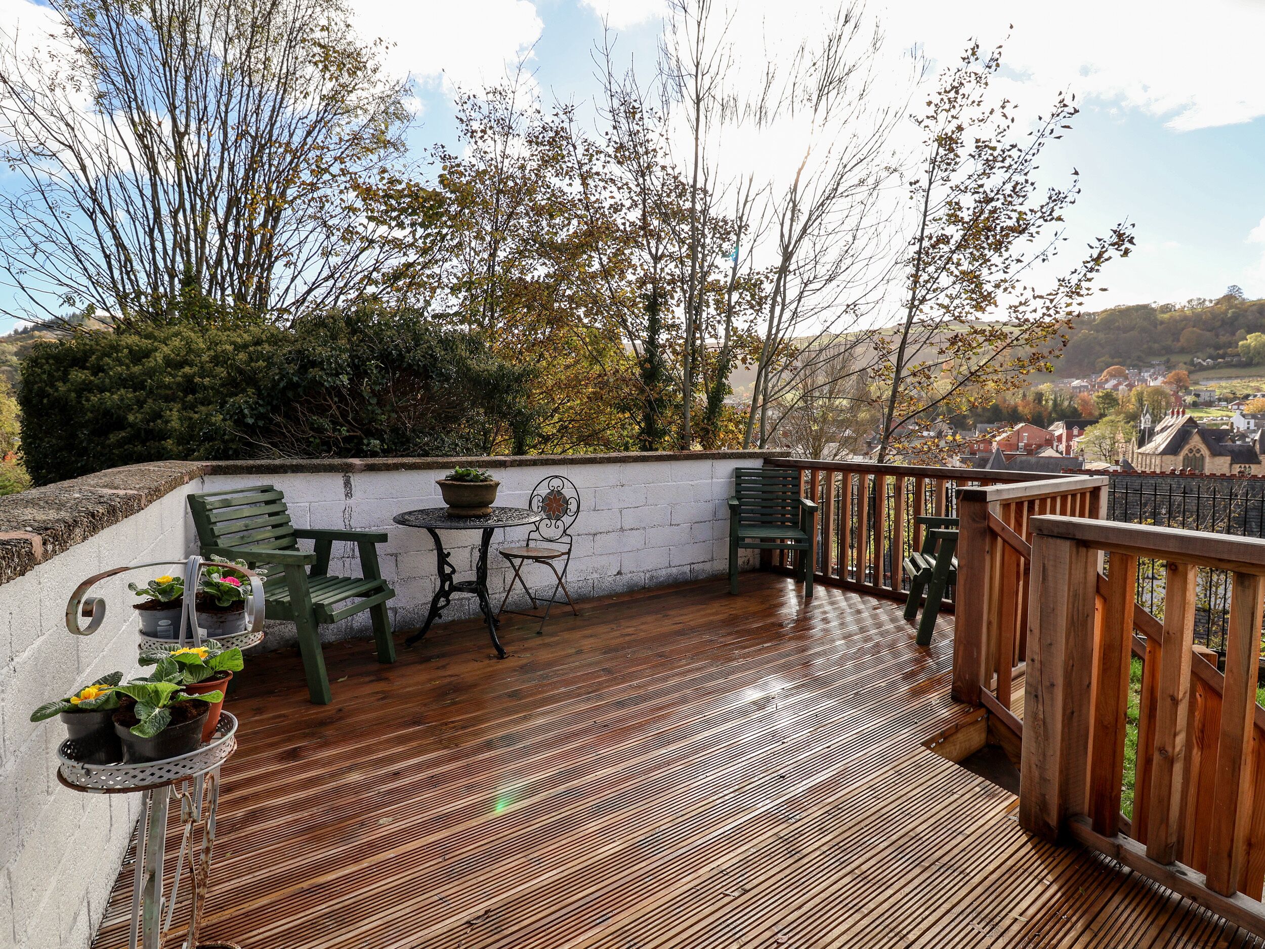 Une terrasse en bois avec une table et des chaises, entourée d’un mur blanc et d’arbres.