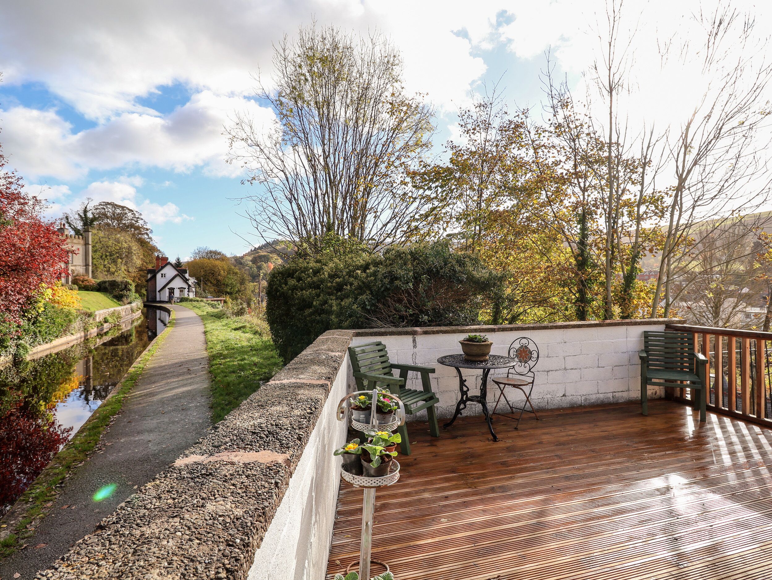 A wooden deck with a table and chairs, a small garden area with potted plants, and a pathway leading to a white building.