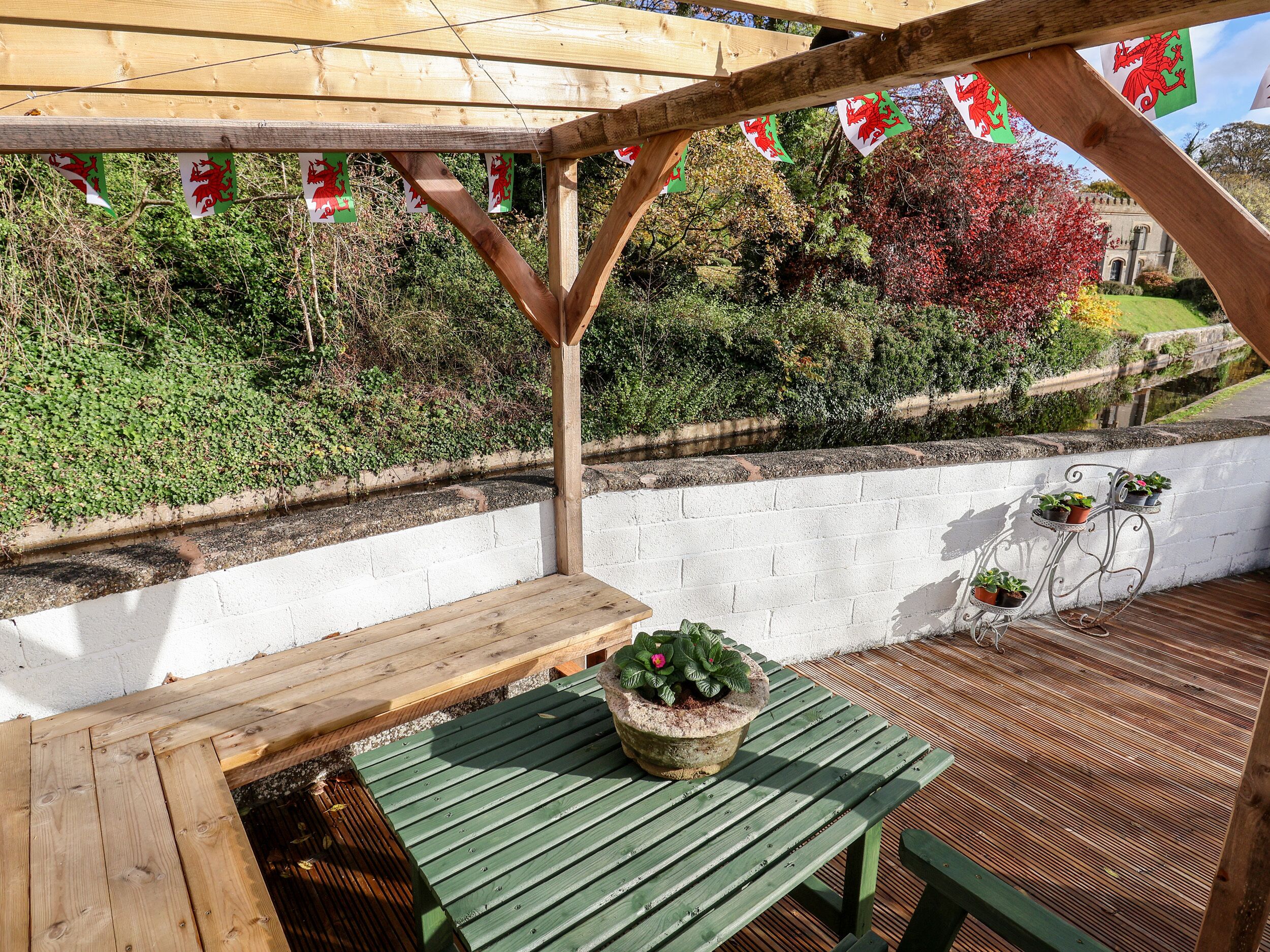 A wooden deck with a bench and potted plants, surrounded by a white wall and greenery.