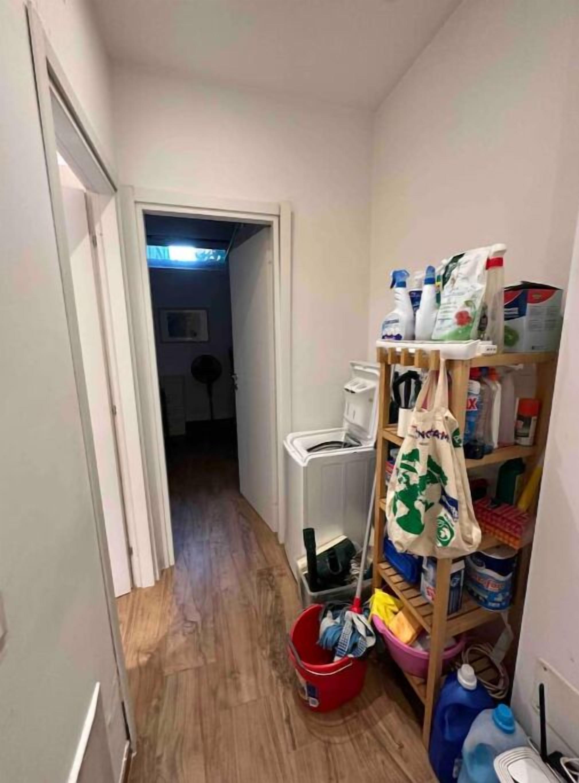 En smal hall med en plats för tvätt, inklusive en tvättmaskin, en röd hink och rengöringsmedel.