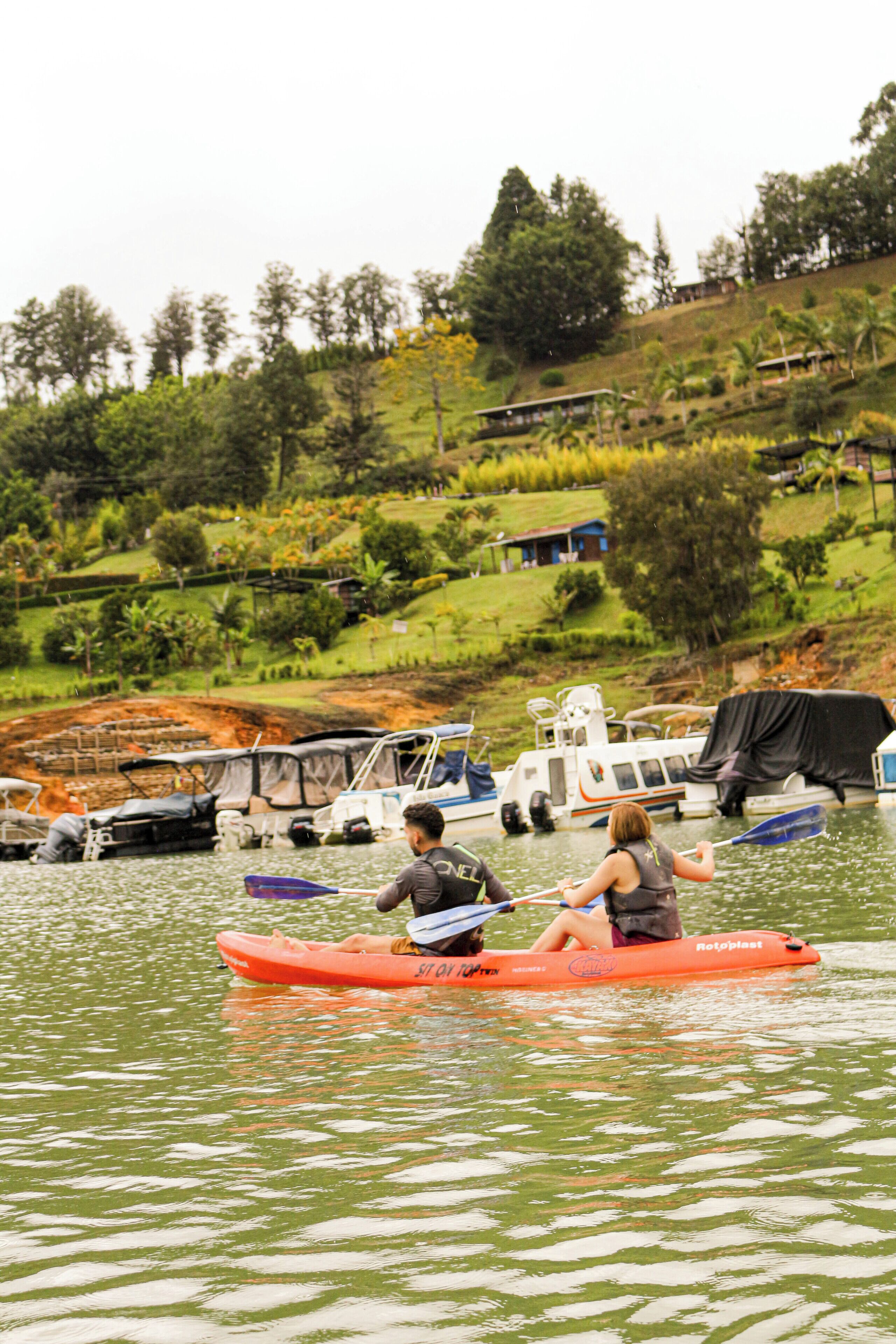 A couple kayaking on a calm river with boats docked nearby.
