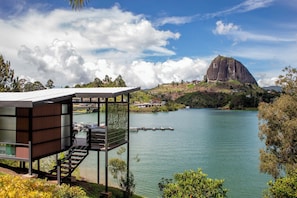 Un edificio moderno con fachada de vidrio que da a un lago, una destacada formación rocosa al fondo y cielo despejado.