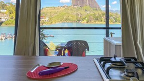 A room with a large window overlooking a lake and a prominent rock formation. - Cabañas Navegar (El Peñol)