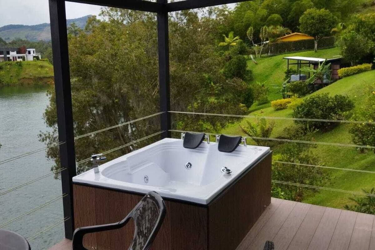 A balcony with a hot tub overlooking a river, surrounded by lush greenery and hills.