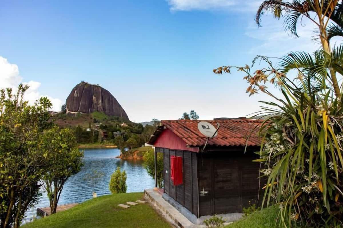 A small wooden building with a red roof near a lake, surrounded by greenery and a large rock formation in the background.