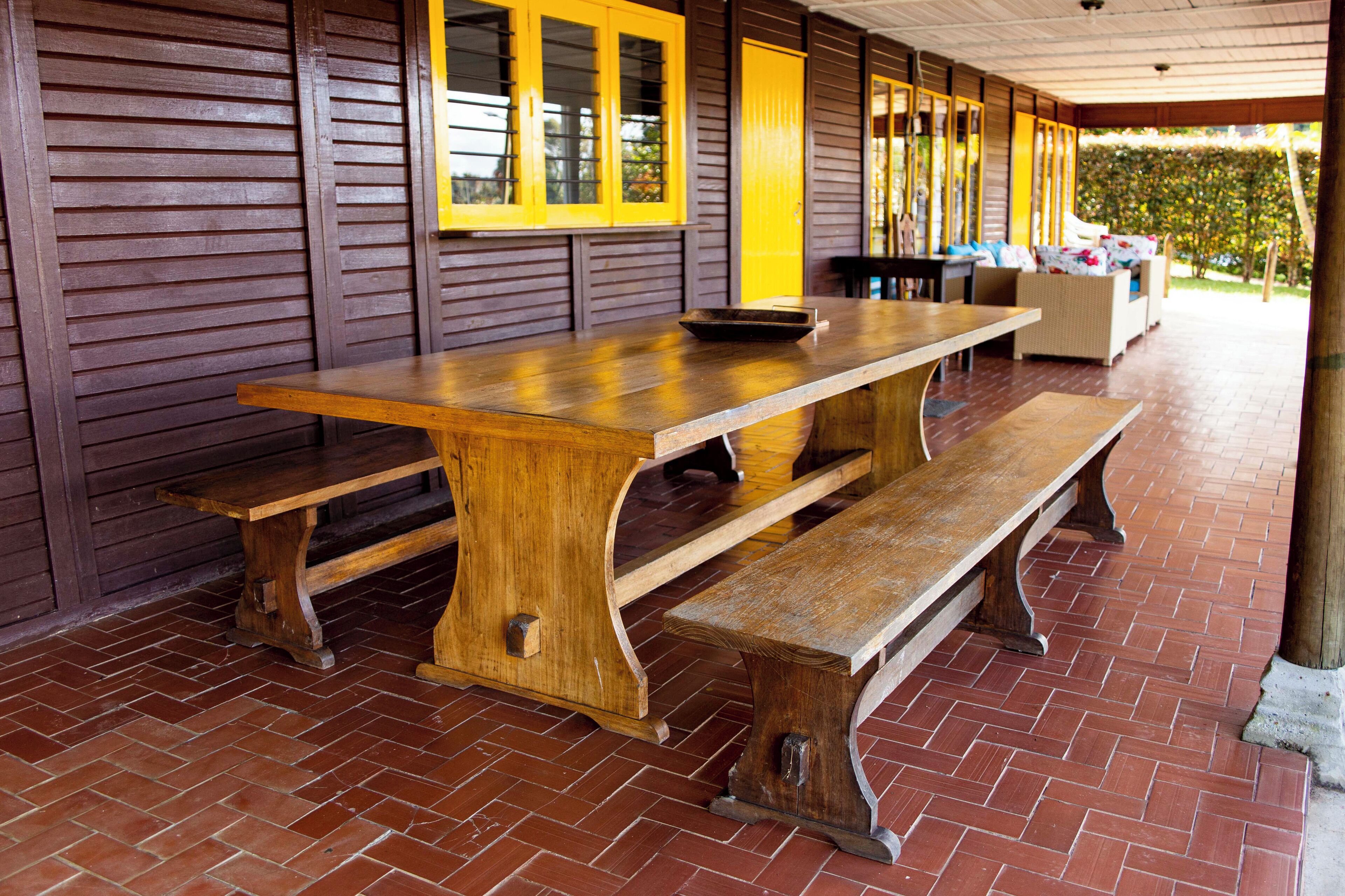 Um alpendre coberto com mesa e bancos de madeira, piso de tijolos e uma porta amarela.