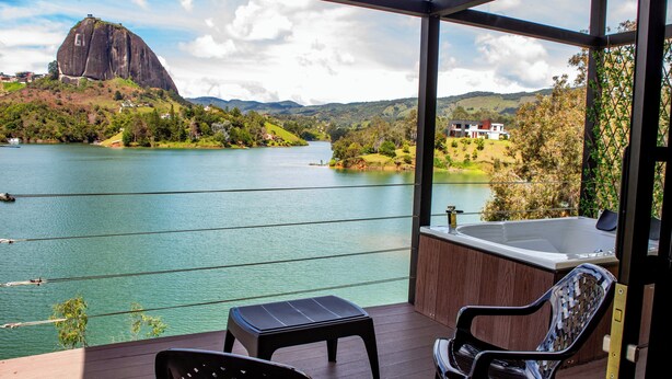 A balcony with two chairs and a small table, overlooking a lake with a large rock formation and houses in the background. - Cabañas Navegar (El Peñol)