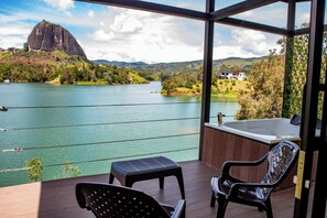 A balcony with two chairs and a small table, overlooking a lake with a large rock formation and houses in the background. - Cabañas Navegar (El Peñol)