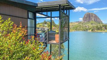 A modern building with a balcony overlooking a lake, a mountain in the background, and lush greenery in the foreground.