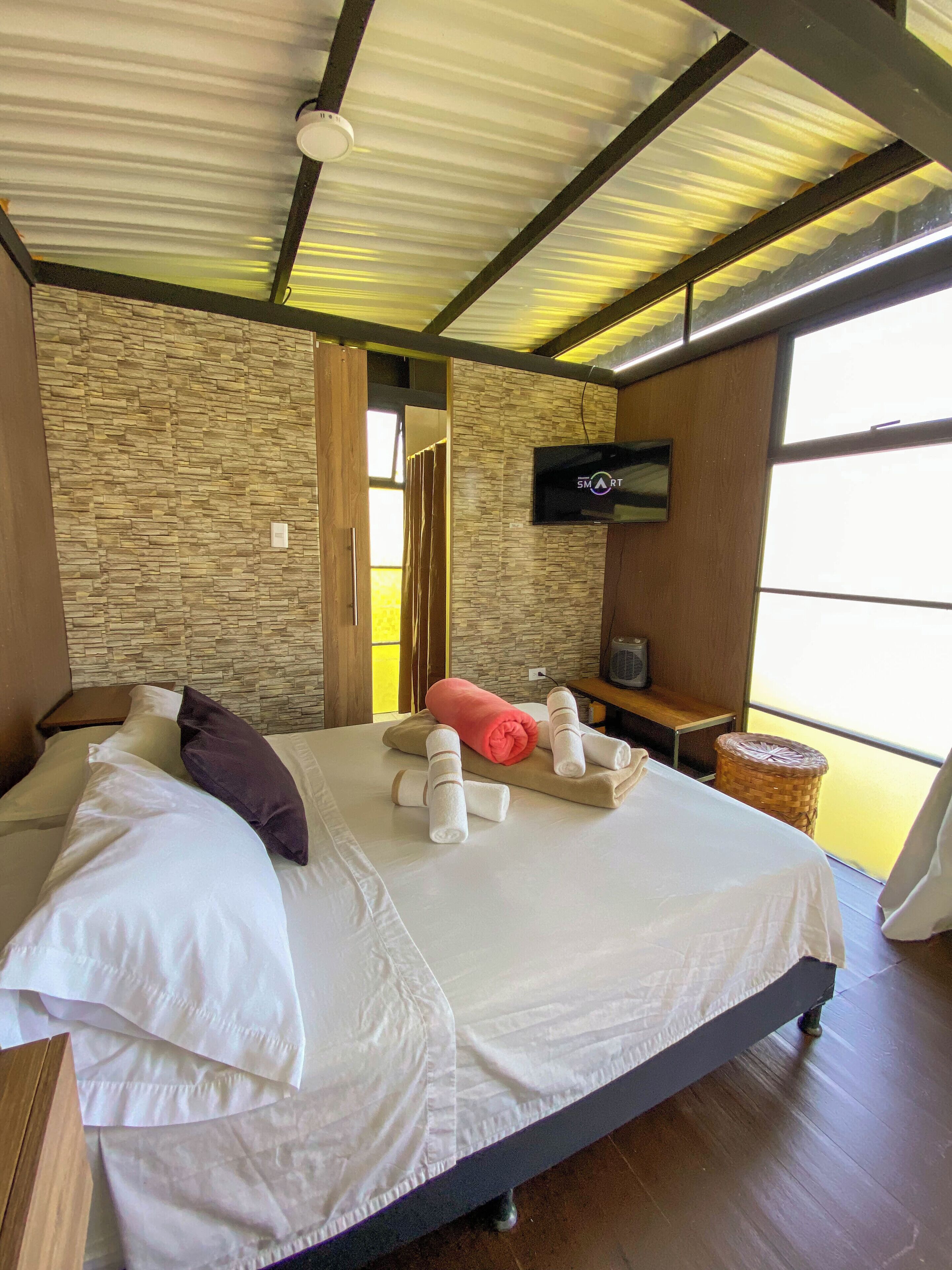 Quarto com parede de pedra, cama com roupa de cama branca, televisão na parede e janela.