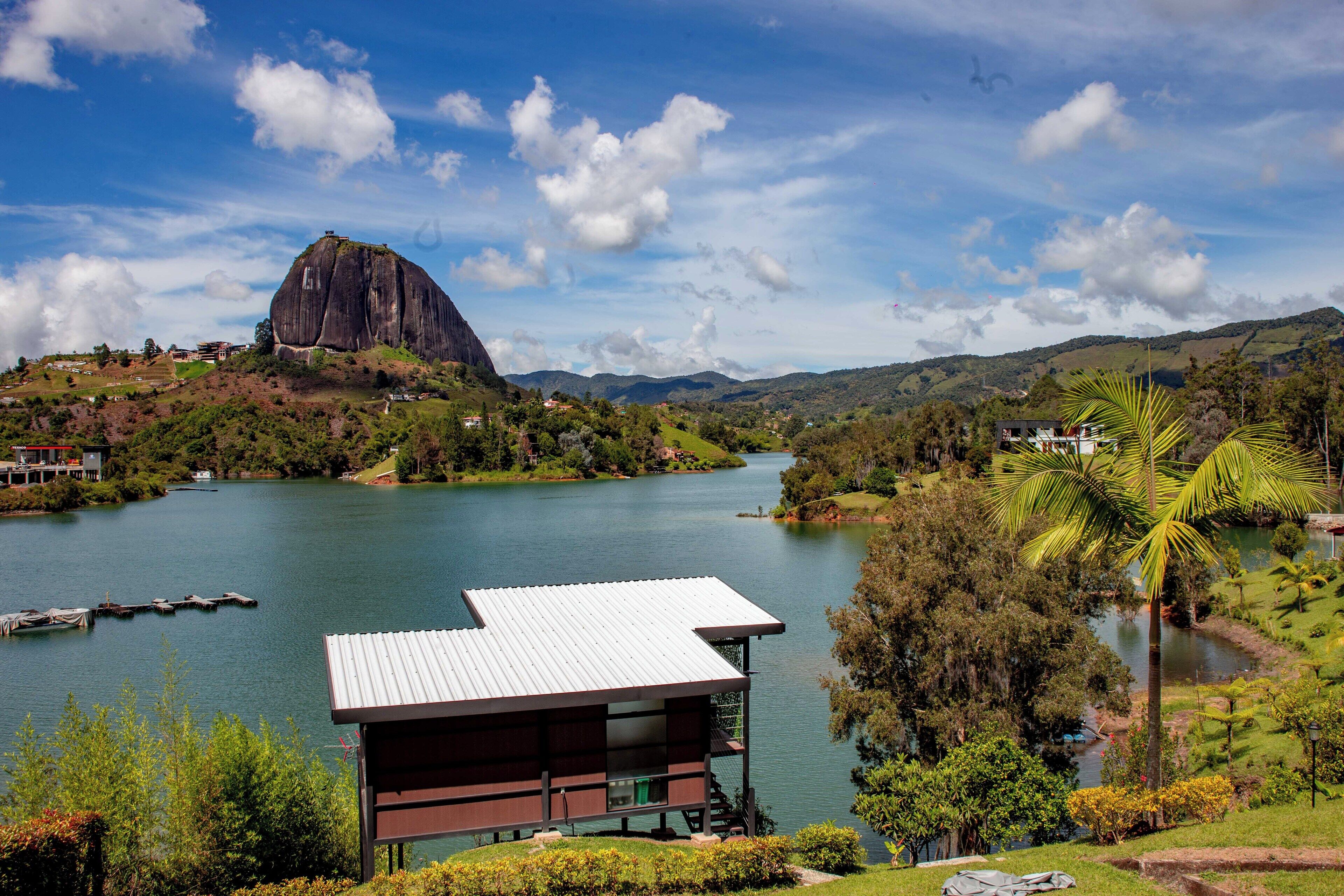 Vista de un lago con una destacada formación rocosa, un pequeño edificio de techo blanco y abundante vegetación circundante.