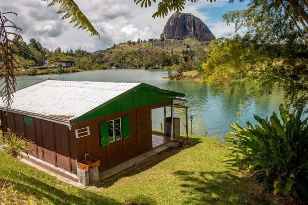 A small cabin with a green roof and a porch, situated by a lake with a large rock formation in the background.