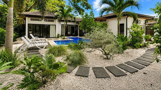 A modern house with a swimming pool, surrounded by tropical vegetation and a gravel pathway.