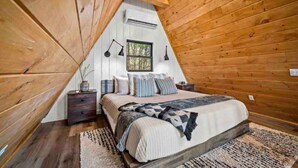 A cozy attic bedroom with a wooden sloped ceiling, a large bed with patterned bedding, a nightstand, and a window with a view of trees. - Secluded View! A-Frame Hot Tub and Fire Pit (Bryson City)
