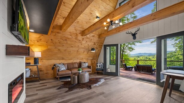 A modern log cabin interior with a wooden ceiling, a leather sofa, a coffee table, and a large window offering a view of the outdoors. - Secluded View! A-Frame Hot Tub and Fire Pit (Bryson City)