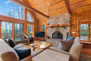 A log cabin interior with a stone fireplace, wooden furniture, and large windows.