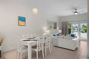 A modern living room with a dining area, white furniture, a ceiling fan, and a view through the glass door.