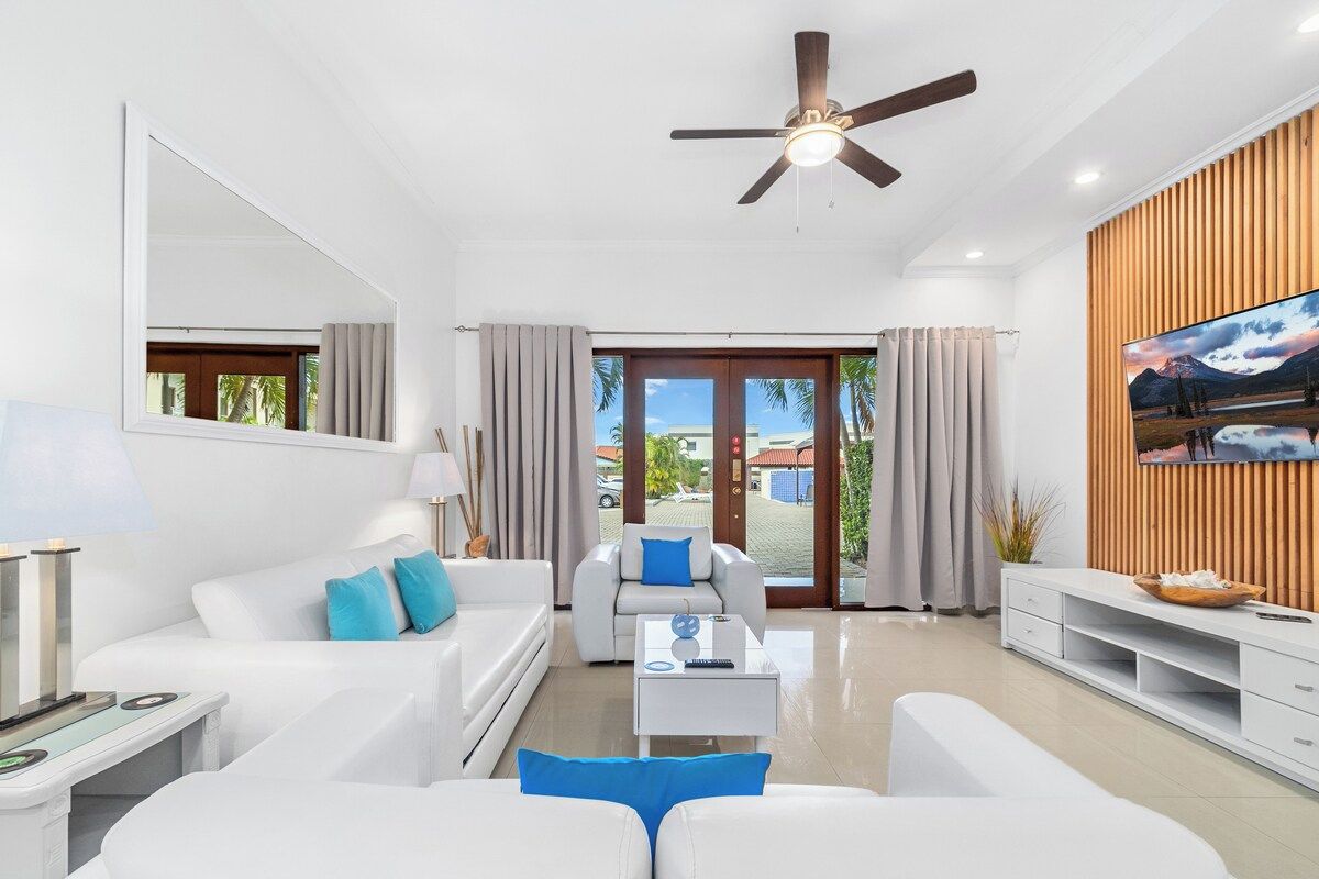 A modern living room with a ceiling fan, white sofas, a coffee table, and a TV on a wooden wall.