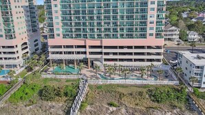 A multi-story hotel with a parking garage, a swimming pool, and a landscaped area with palm trees. - Crescent Keyes 904 - 3BD/3BA Oceanfront Condo (North Myrtle Beach)