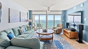 A spacious living room with a large window overlooking the sea, a ceiling fan, a round coffee table, and a flat-screen TV. - Crescent Keyes 904 - 3BD/3BA Oceanfront Condo (North Myrtle Beach)