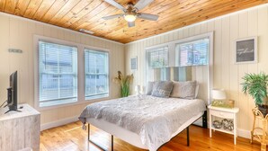 A bedroom with a wooden ceiling, large windows with blinds, a bed with a gray bedspread, a bedside table with a lamp, and a small plant. - Pet Friendly 3 BR House - Latitude Adjustment (North Myrtle Beach)