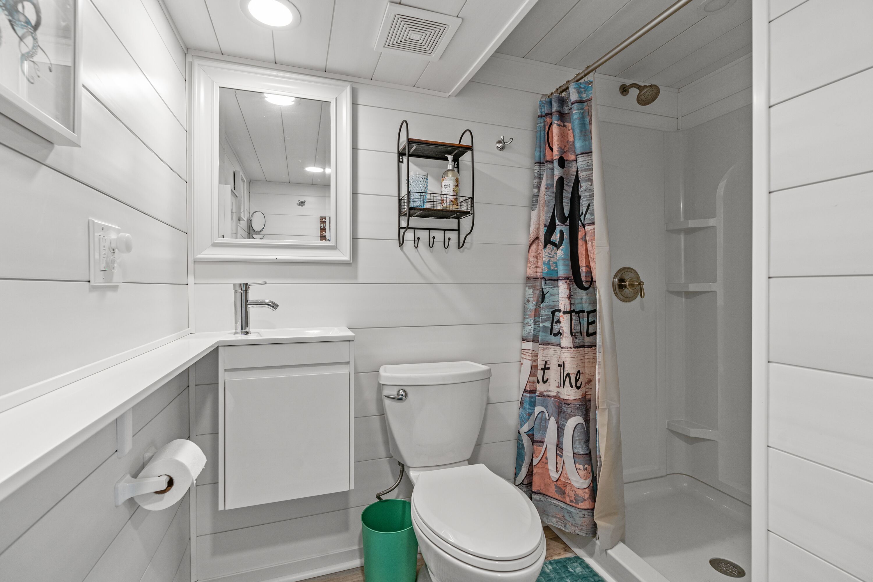 Une salle de bain équipée de toilettes, d’un lavabo, d’un miroir, d’une douche et d’une poubelle verte.
