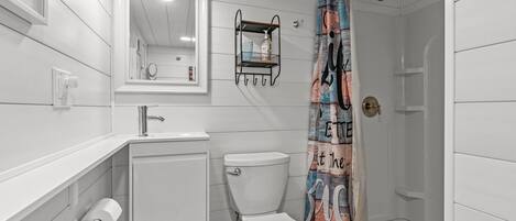 Une salle de bain équipée de toilettes, d’un lavabo, d’un miroir, d’une douche et d’une poubelle verte.