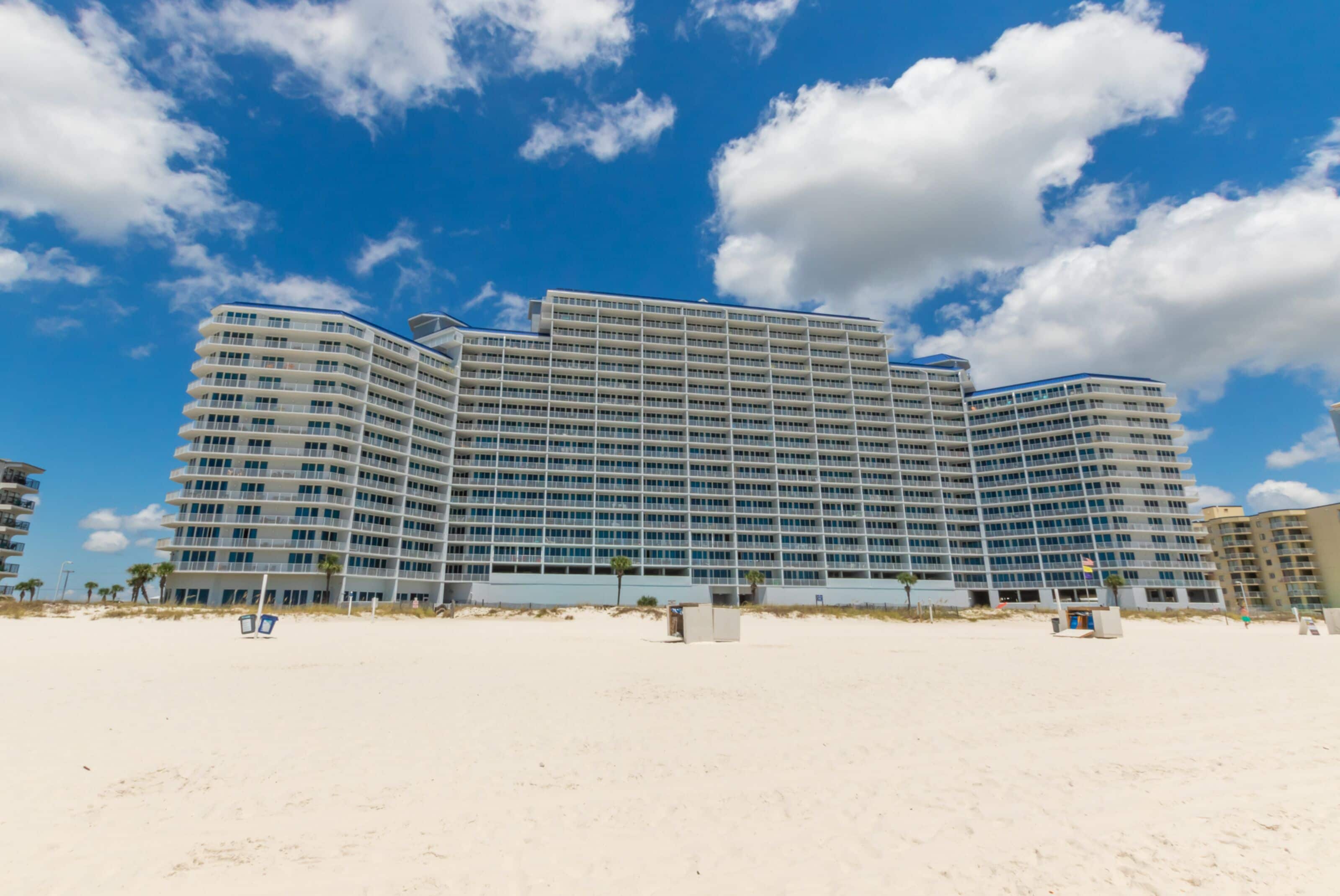 A large, multi-story hotel with a curved facade, located on a beach with clear skies.