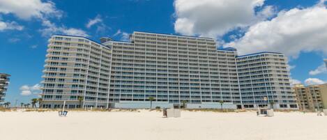 A large, multi-story hotel with a curved facade, located on a beach with clear skies.