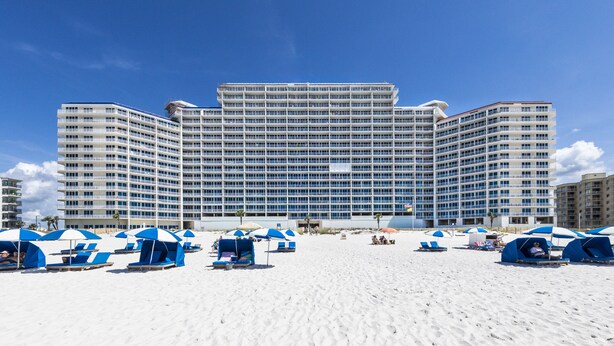 A large hotel with multiple floors and balconies, a sandy beach in front, and several beach umbrellas and lounge chairs. - Lighthouse 1004 (Gulf Shores)