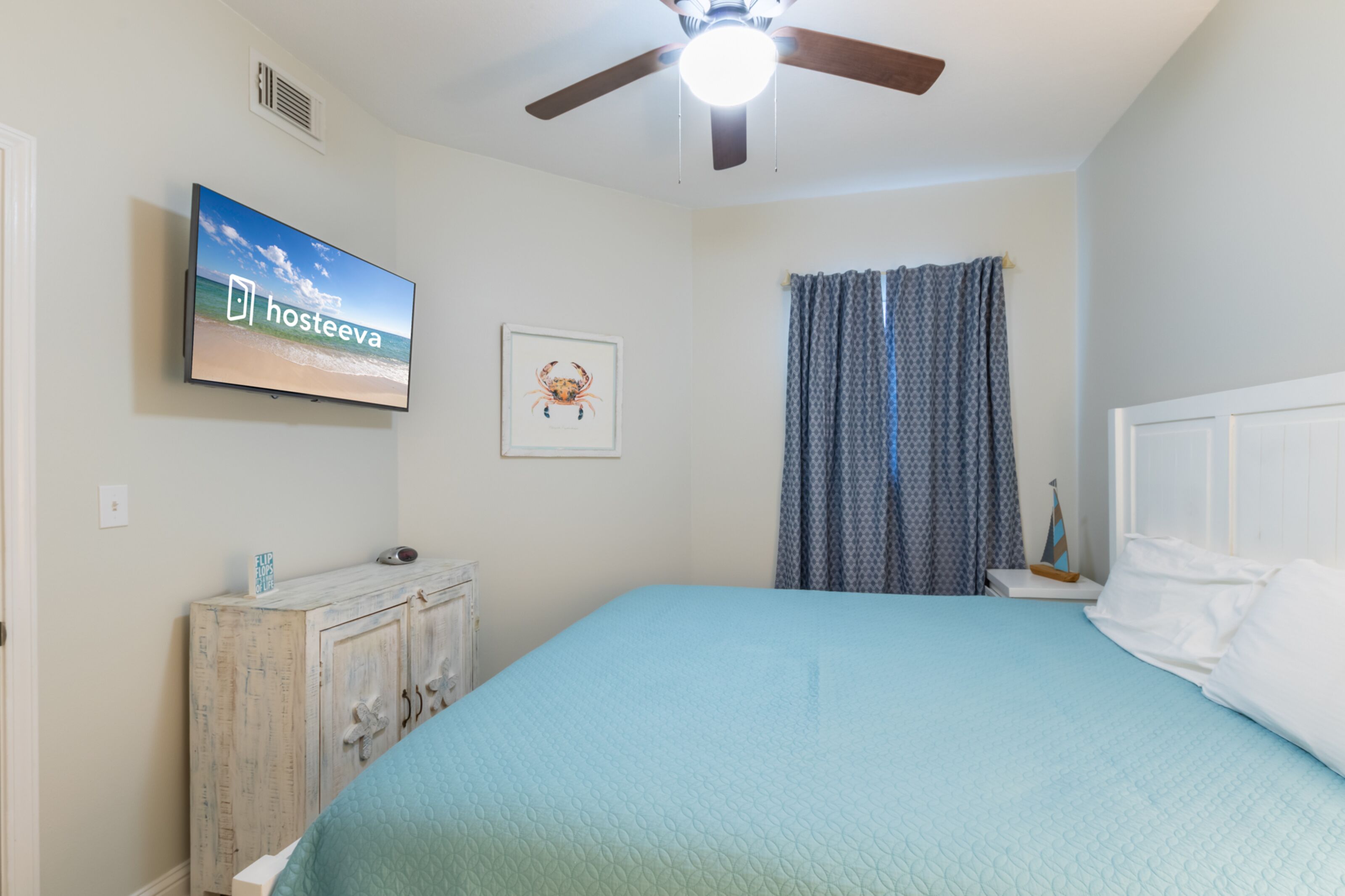 Une chambre à coucher avec un lit, un ventilateur de plafond, un téléviseur affichant le logo « hosteeva », un tableau encadré et une commode.