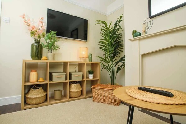 A living room with a TV, shelves with decorative items, a large plant, and a round table with a remote.